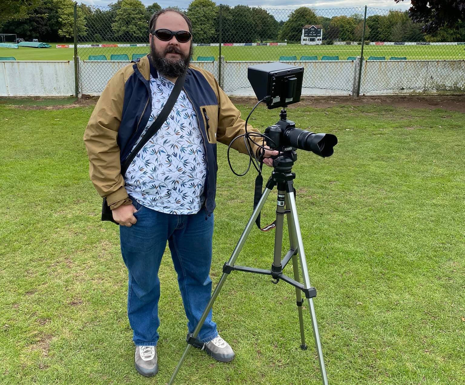 Steve, an SVI member is pictured standing with the hand on his camera and tripod in front of a cricket pitch. It's clear the camera has adjustments to help take photographs with a visual impairment.
