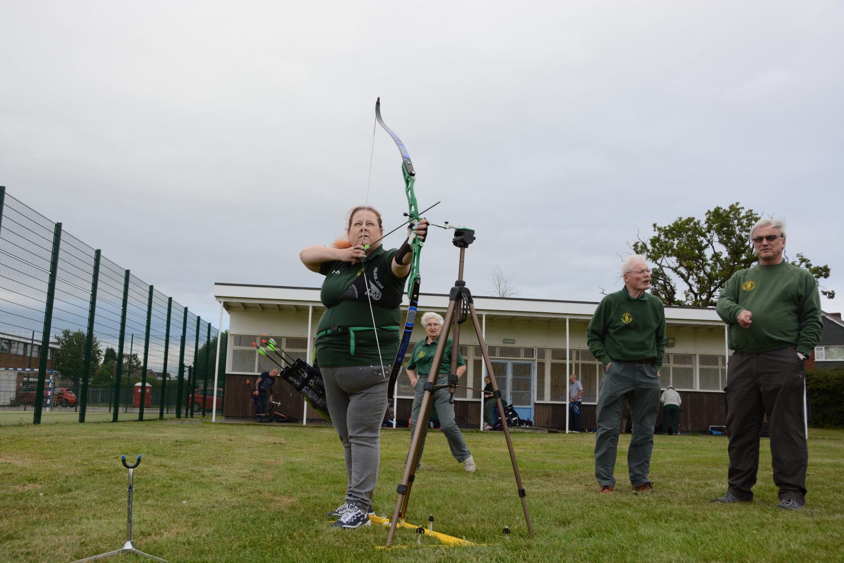 Photograph of a white woman, wearing a green polo top, standing in a field outside a sports club holding a bow and arrow. She draws her arrow to the right of the frame and her look and stance is defiant. The coach and spotter stand behind her.  