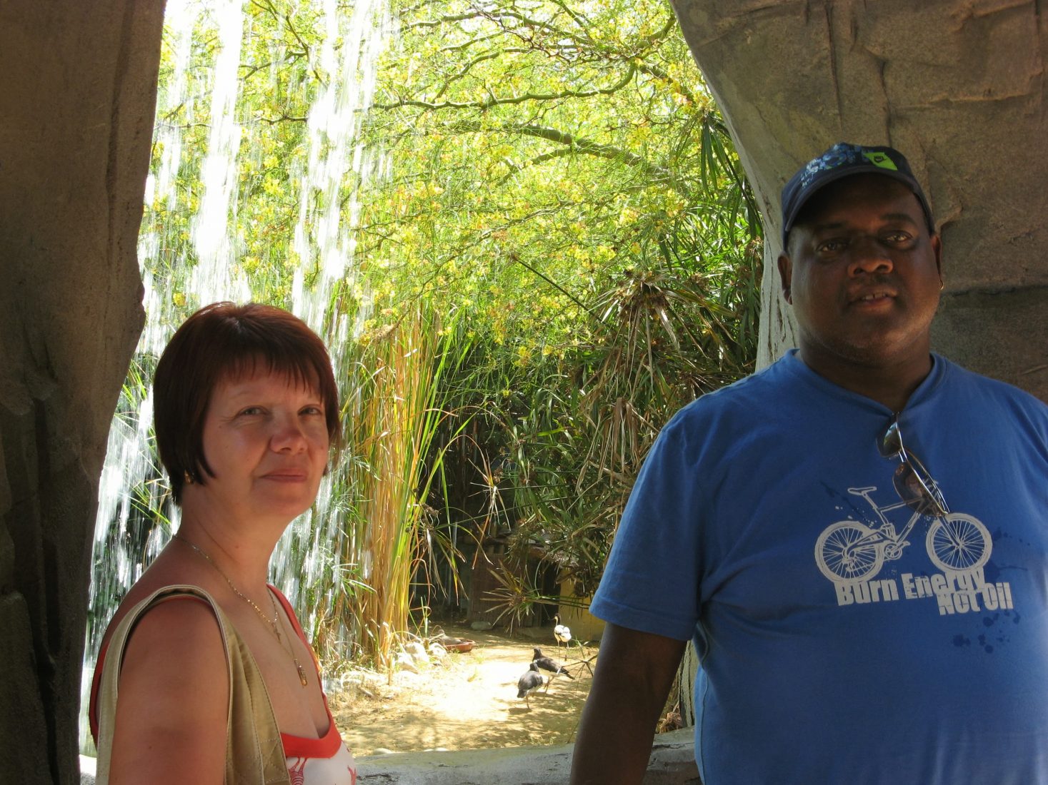 A white woman with short reddish hair and a black man wearing a blue t-shirt face the camera. In the background the sunlight catches on the leaves of the trees. 