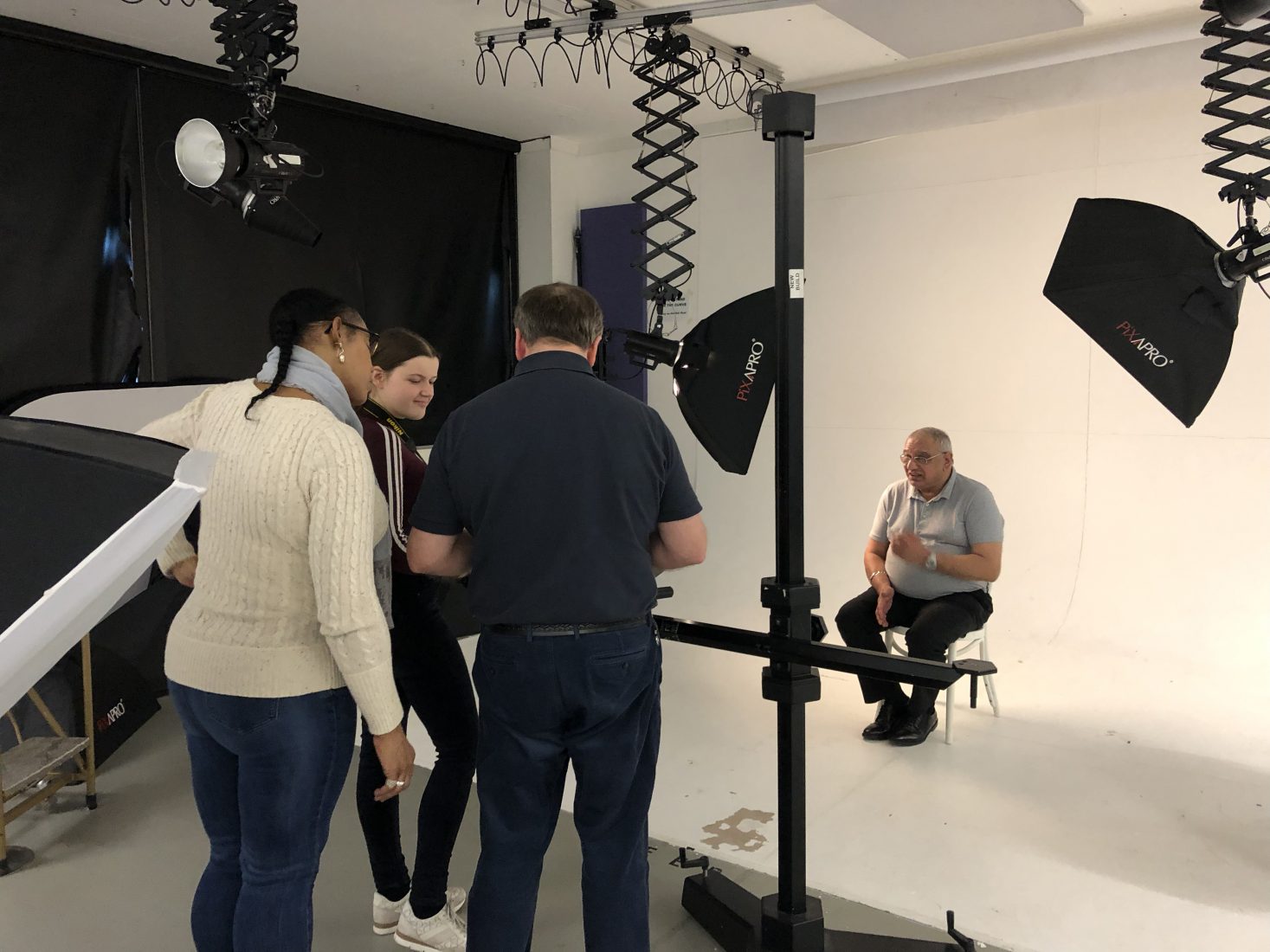 A man sits in a white studio which is lit up by lights. Three people, two women and a man are grouped together as if preparing to photograph the man.