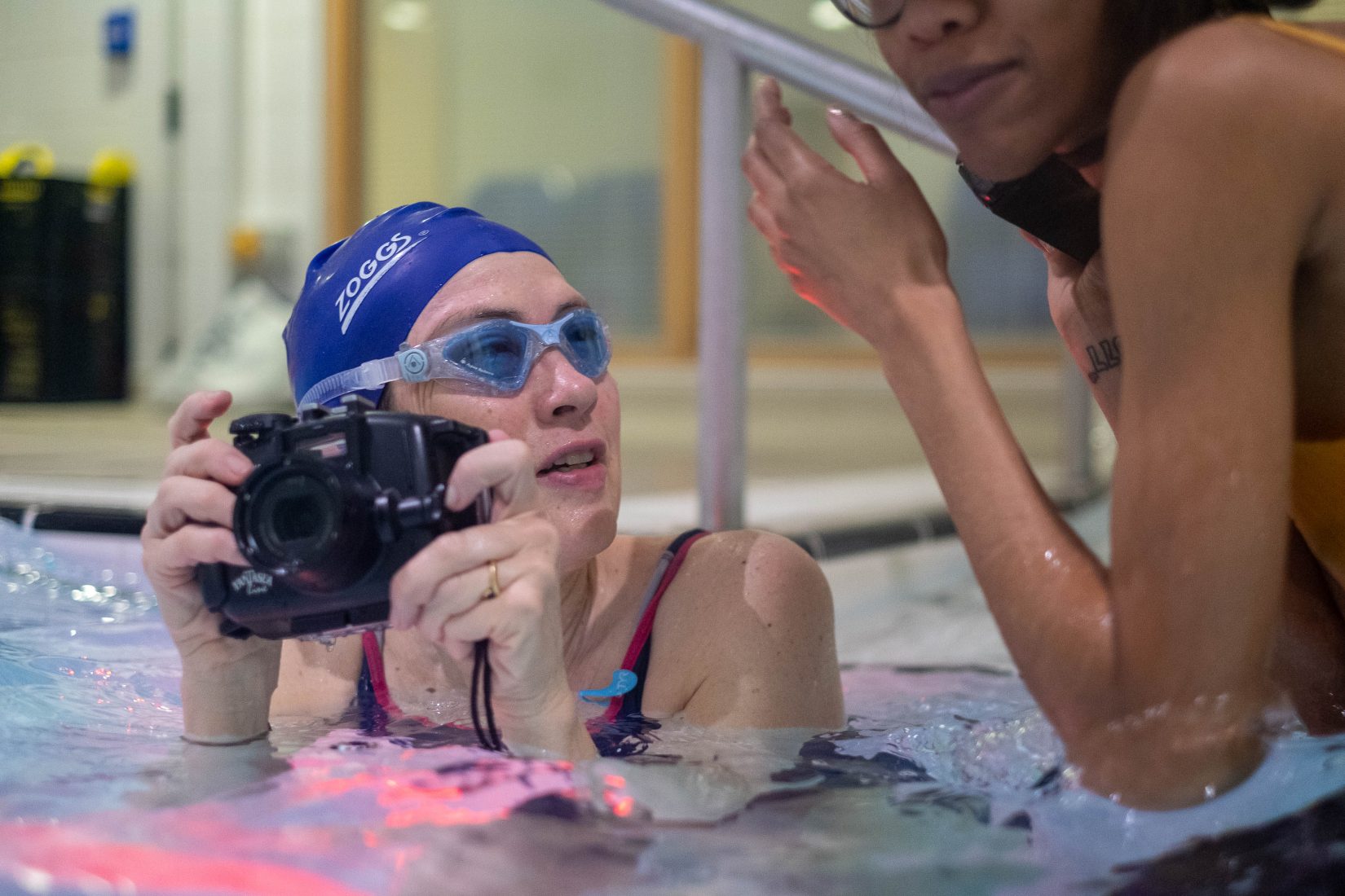 A woman wearing a swimming cap and goggles is in a pool, holding a camera in both hands just above the water. She looks at the woman beside her in the pool.