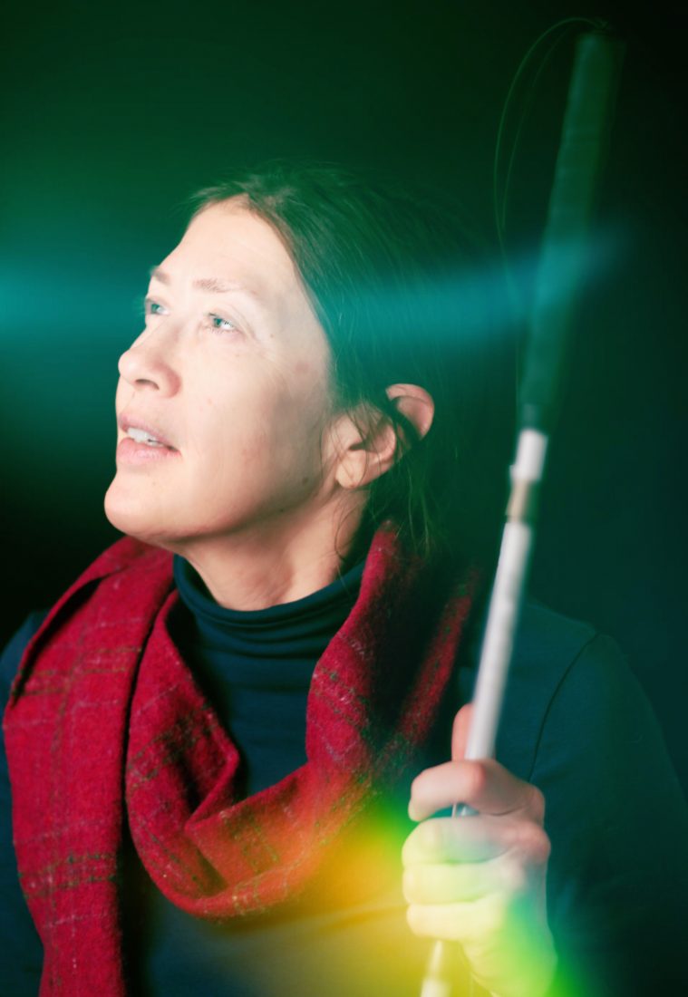 A woman in profile holding a white cane. Bright light flashes across her face, a translucent rainbow stripe across her hand.