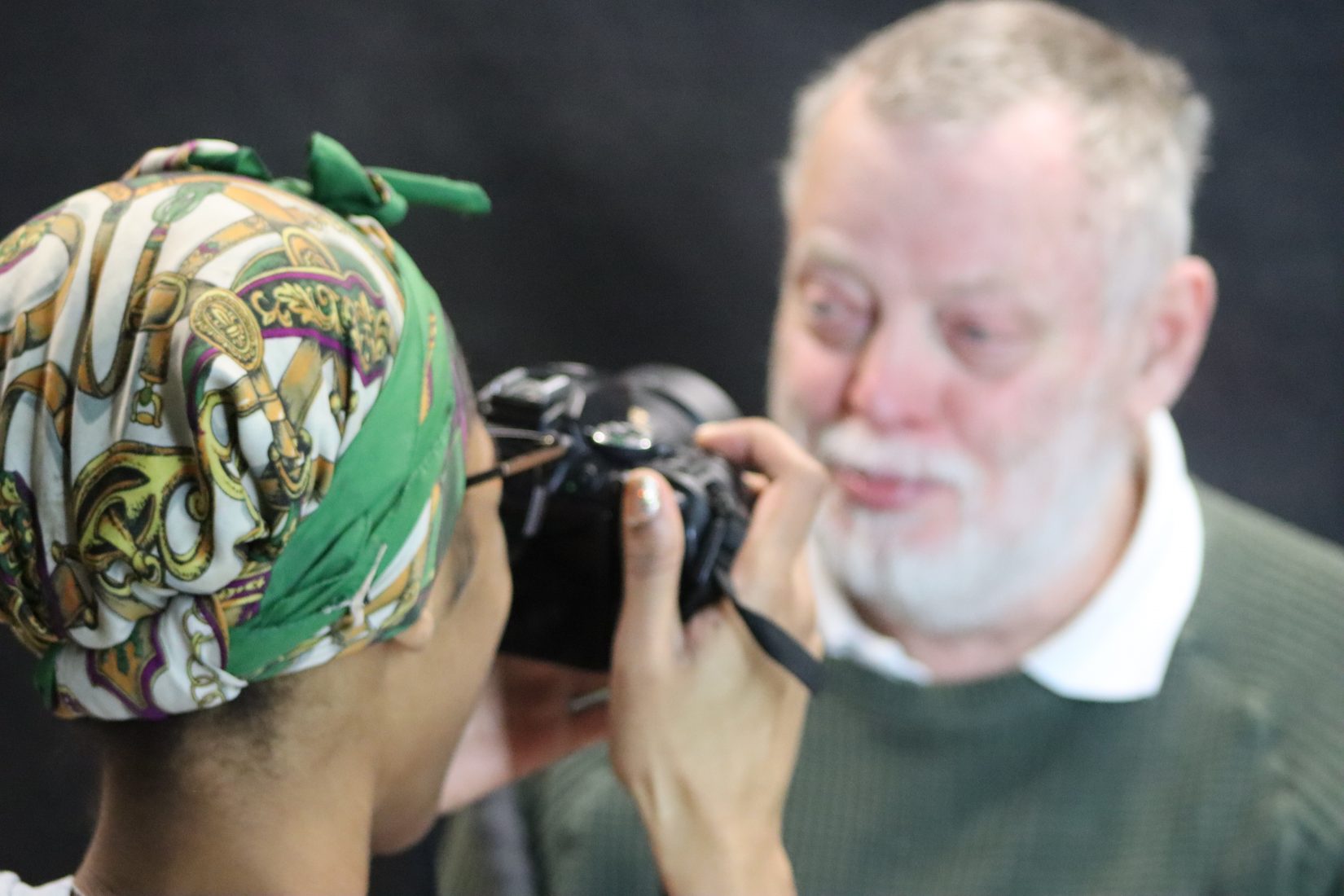 Woman in foreground with a colourful headscarf photographs a man with a grey hair and beard in front of a dark studio backdrop.