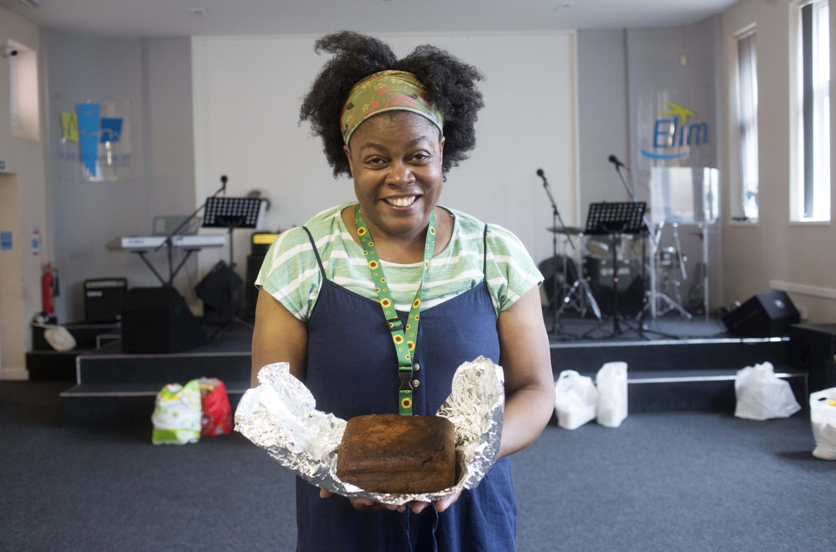 Photograph of a woman with black hair and a green headband, smiling while holding a cake in the foodbank she works at. She’s wearing a stripey green top with a blue dress and has a lanyard on with sunflower fabric.